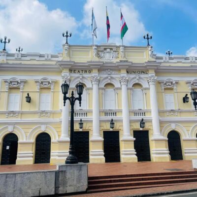 Fachada neoclásica amarilla del Teatro Municipal Enrique Buenaventura en Cali con banderas de Colombia, Cali y Valle del Cauca, y escaleras de acceso.